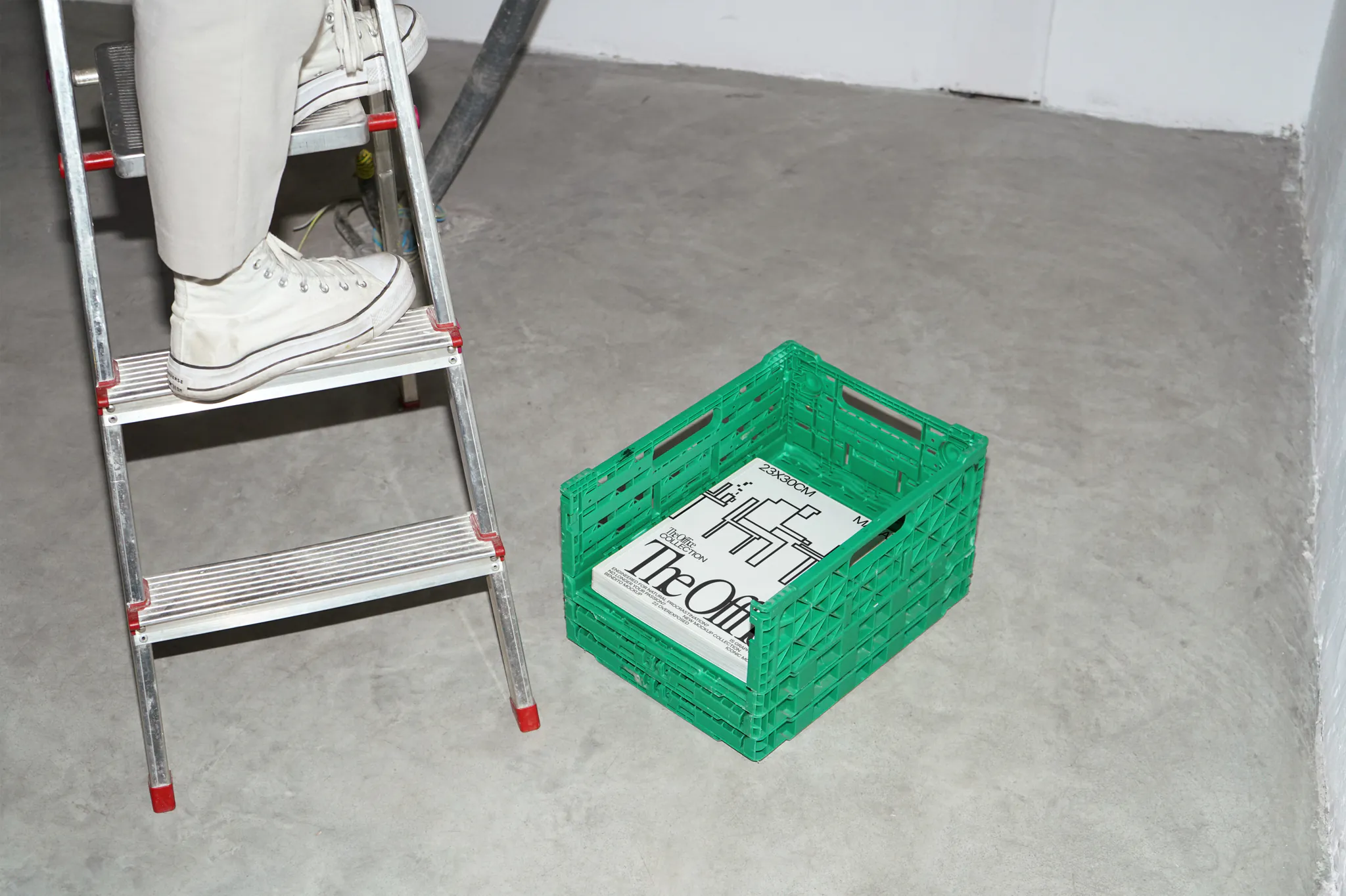 Magazine mockup inside a green plastic box over a concrete floor near a person who is on a ladder