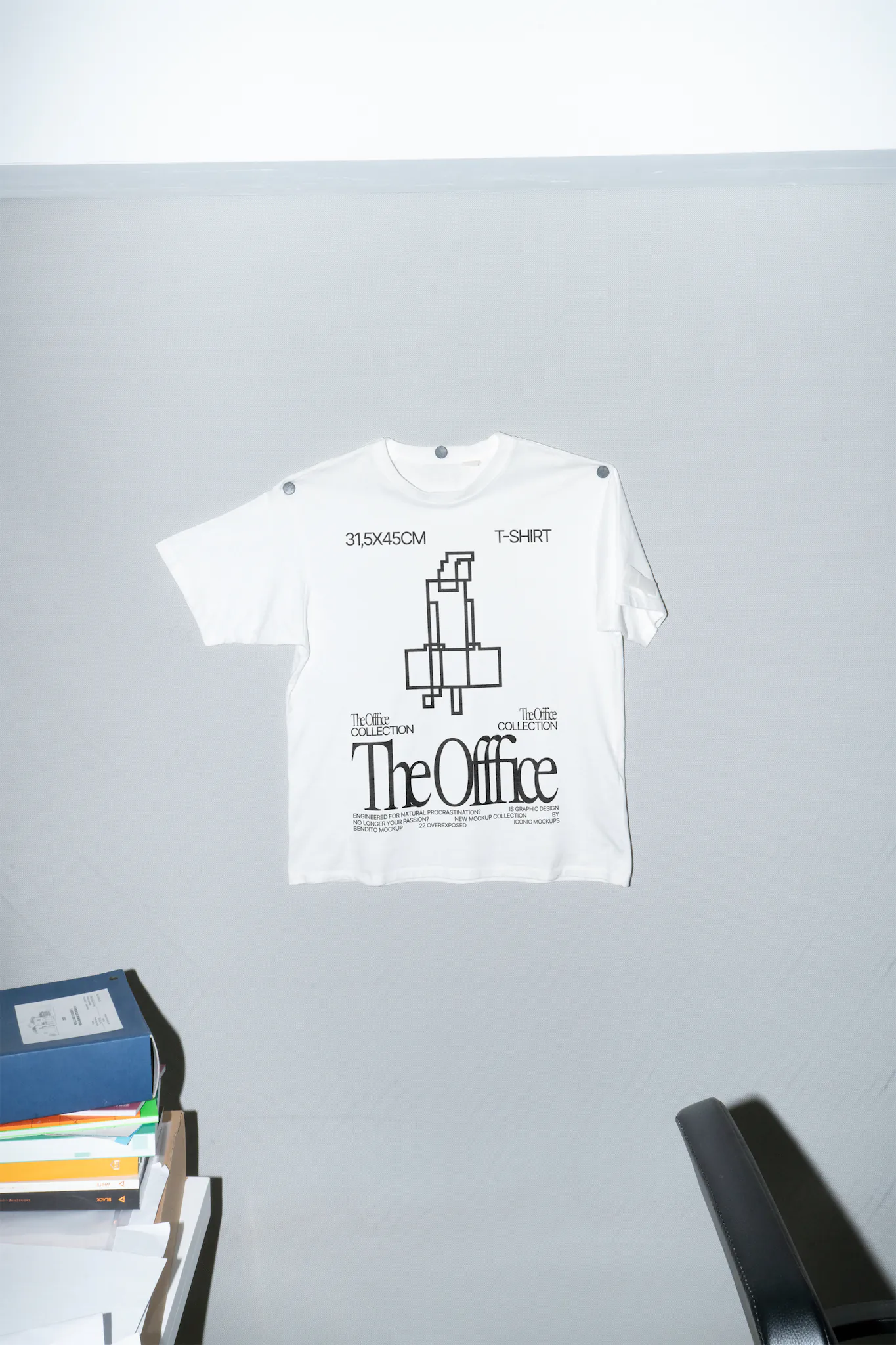 T-shirt mockup hanging on a wall with one of its sleeves open and the other one closed next to an office table with papers on top