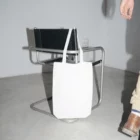 Tote bag mockup hanging from a black leather chair over a concrete floor with white walls around and a man walking near the scene