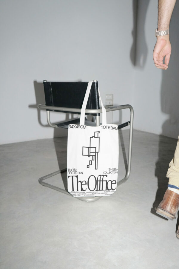 Tote bag mockup hanging from a black leather chair over a concrete floor with white walls around and a man walking near the scene