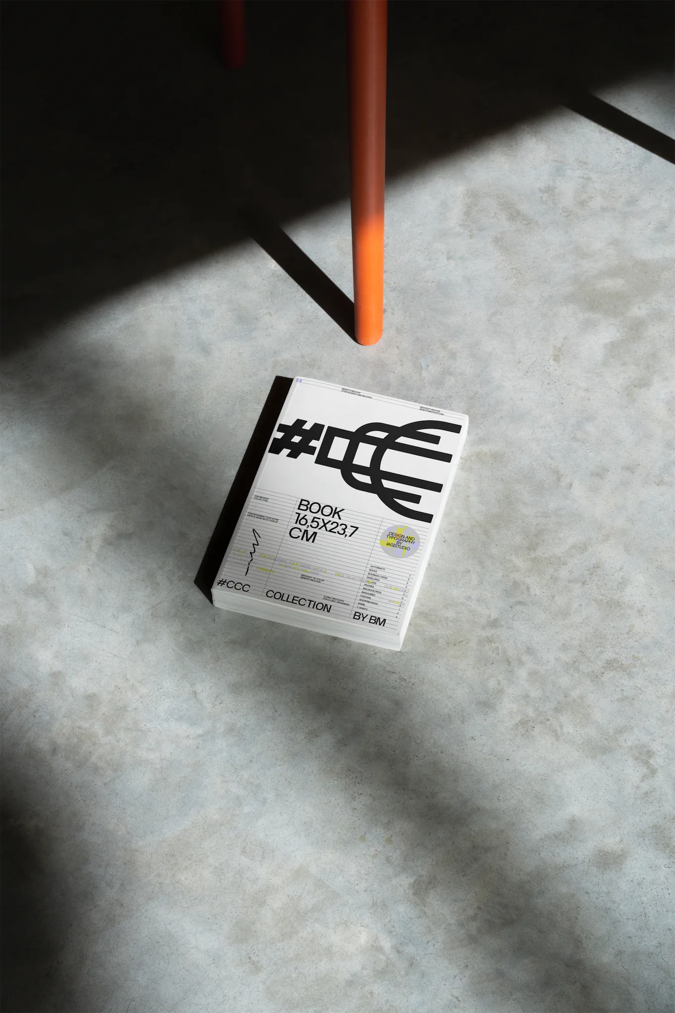 Book format mockup over an industrial floor and placed next to an orange chair. Editorial mockup.