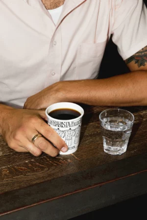Hand holding white coffee cup mockup in a coffee shop. Coffee cup mockup being grabbed by a man, wearing a beige t-shirt. Hand holding cardboard coffee cup mockup, paper cup mockup, tea cup mockup, high quality mockup, PSD file.