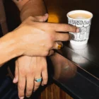 Hand holding a disposable coffee cup mockup laying on a metallic counter. Coffee cup mockup being grabbed by a man in a cafeteria, wearing a beige t-shirt. Hand holding cardboard coffee cup mockup, paper cup mockup, tea cup mockup, high quality mockup, PSD file.