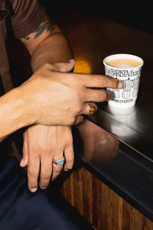 Hand holding a disposable coffee cup mockup laying on a metallic counter. Coffee cup mockup being grabbed by a man in a cafeteria, wearing a beige t-shirt. Hand holding cardboard coffee cup mockup, paper cup mockup, tea cup mockup, high quality mockup, PSD file.