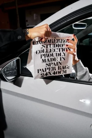 Paper bag mockup grabbed by a person who is giving it to another person who is inside a white car in the parking of a burger restaurant. Print mockup. Paper bag PSD file. Takeaway mockup. Fast food mockup. Premium quality mockup. Burger PSD high-quality editable file.
