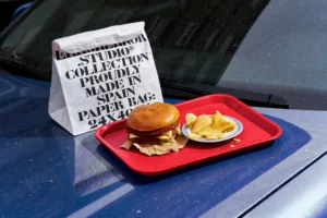 Paper bag mockup on top of the hood of a blue car in the parking of a burger restaurant next to a red tray with burger menu with fries. Bag mockup. Paper bag PSD file. Takeaway mockup. Fast food mockup. High-quality paper bag mockup. Takeaway PSD high-quality editable file.