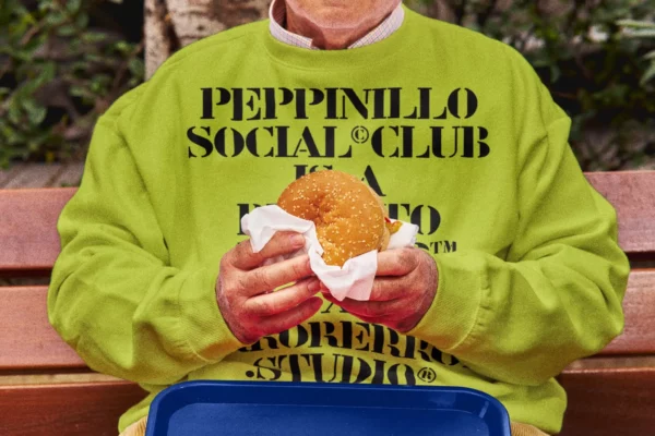 Sweatshirt mockup worn by an old person who is eating a burger sitting outside on the bench of a burger restaurant. Clothing mockup. Jumper mockup. Takeaway PSD file. Takeaway mockup. Fast food mockup. Premium quality sweatshirt mockup. Takeaway PSD high-quality editable file. Burger high-quality mockup.