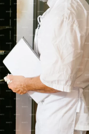 A professional baker holding a notebook in a bakery kitchen setting.
