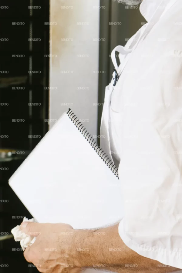 Zoom in of a professional baker holding a notebook in a bakery kitchen setting.