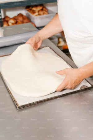 Hands stretching fresh bread dough on a baking tray in a kitchen.