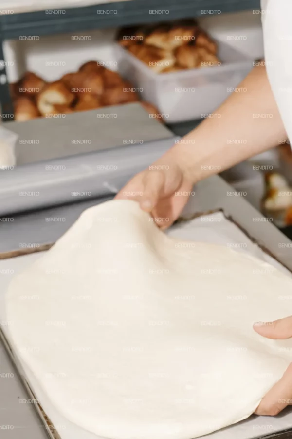 Zoom in of hands stretching fresh bread dough on a baking tray in a kitchen.