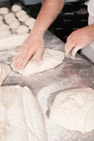Hands kneading bread dough on a floured countertop in a bakery setting.