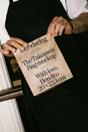 Mockup of person holding a brown paper bag while wearing a black apron. Perfect for eco-conscious food branding.