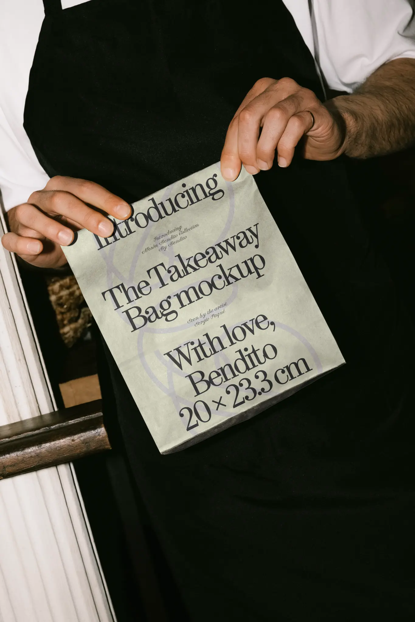 Color Version: Mockup of person holding a brown paper bag while wearing a black apron. Perfect for eco-conscious food branding.