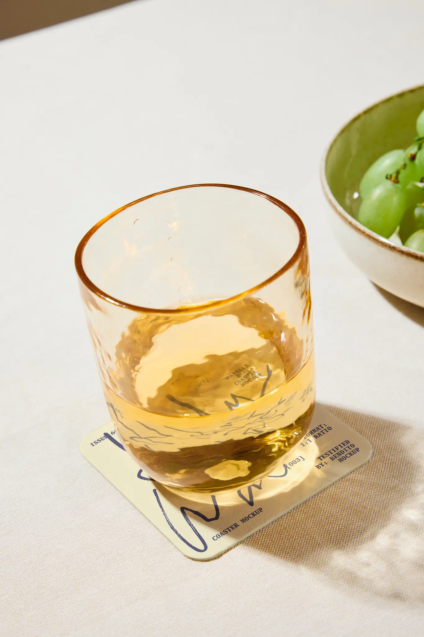 A glass of amber-colored liquid sits on a printed coaster on a light-colored table next to a green bowl filled with green grapes.