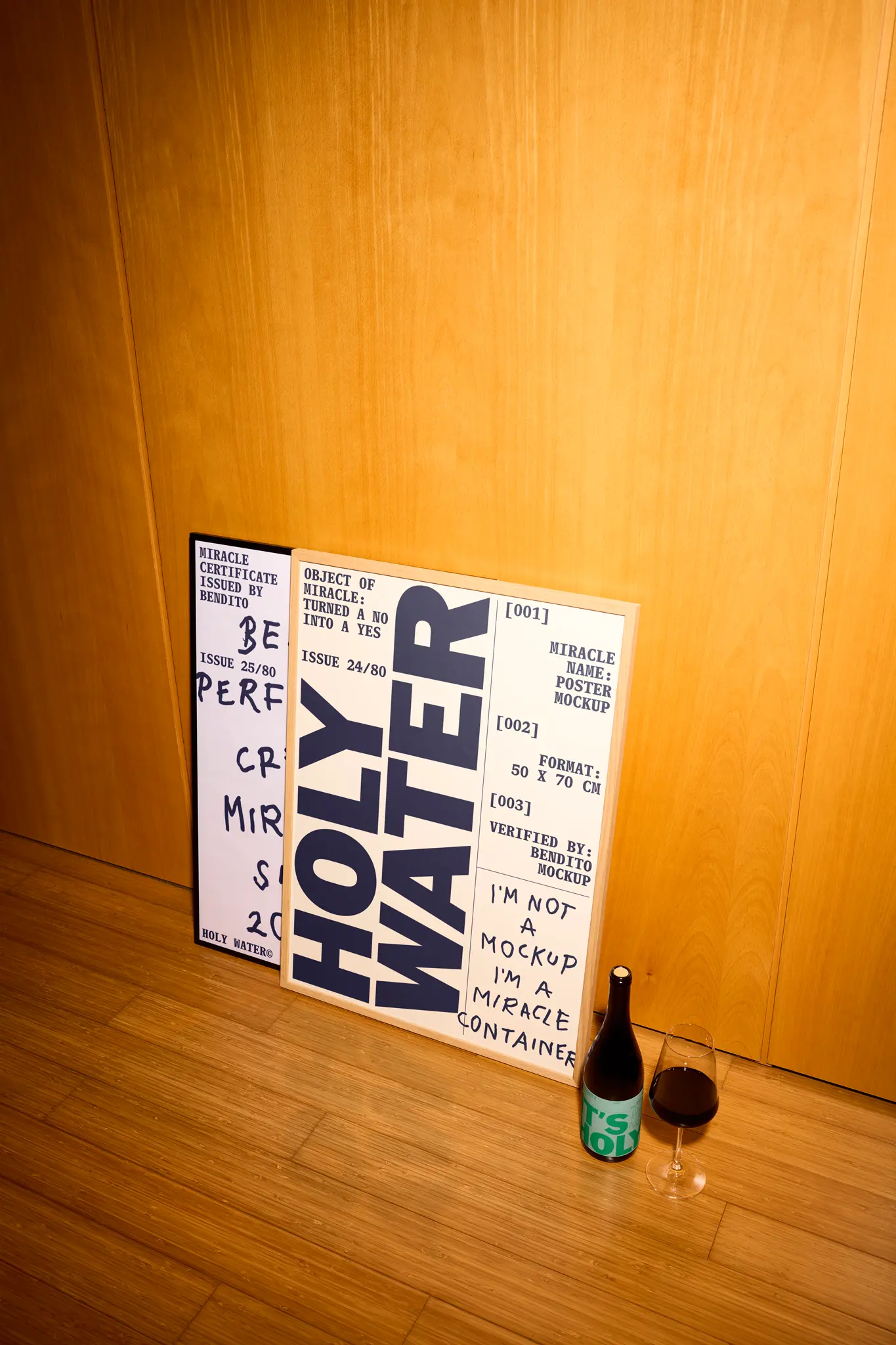 A framed print reading HOLY WATER in bold letters leans against a wooden wall on a wood floor. Next to it are a wine bottle and a filled wine glass.