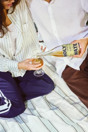 A person pours a beverage labeled Holy Water into a glass held by another person as they sit on a striped blanket, both dressed in casual, comfortable clothes—perfect for a product mockup scene.