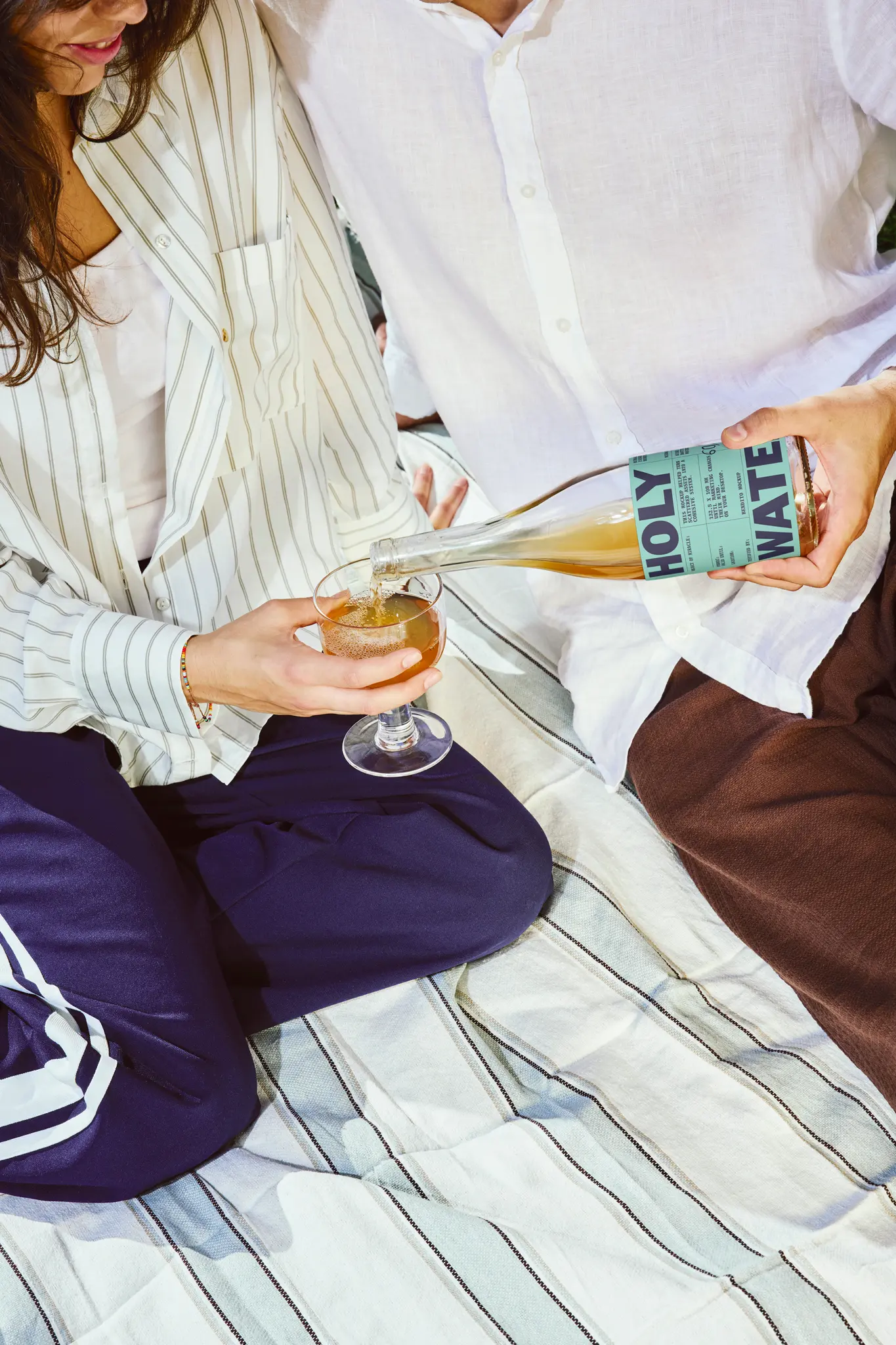 Two people sit on a striped blanket in a relaxed mockup scene, one pouring a drink from a bottle labeled Holy Water into the others glass. Both are dressed casually.