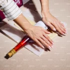 A stock photo shows a person wrapping a bottle of wine in white tissue paper on a beige surface, both hands smoothing the paper. The wine bottle has a red foil top and there is a book partially visible nearby.