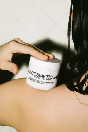 Mockup of a white cosmetic jar (110 × 48 mm) held against a shoulder, captured in a steamy, minimal shower scene. By Bendito Mockup.