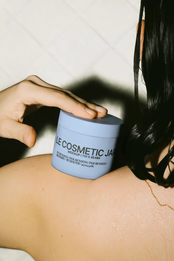 Cosmetic jar mockup in use, placed on a person’s shoulder in a shower setting. Great for skincare or wellness branding at 110 × 48 mm.