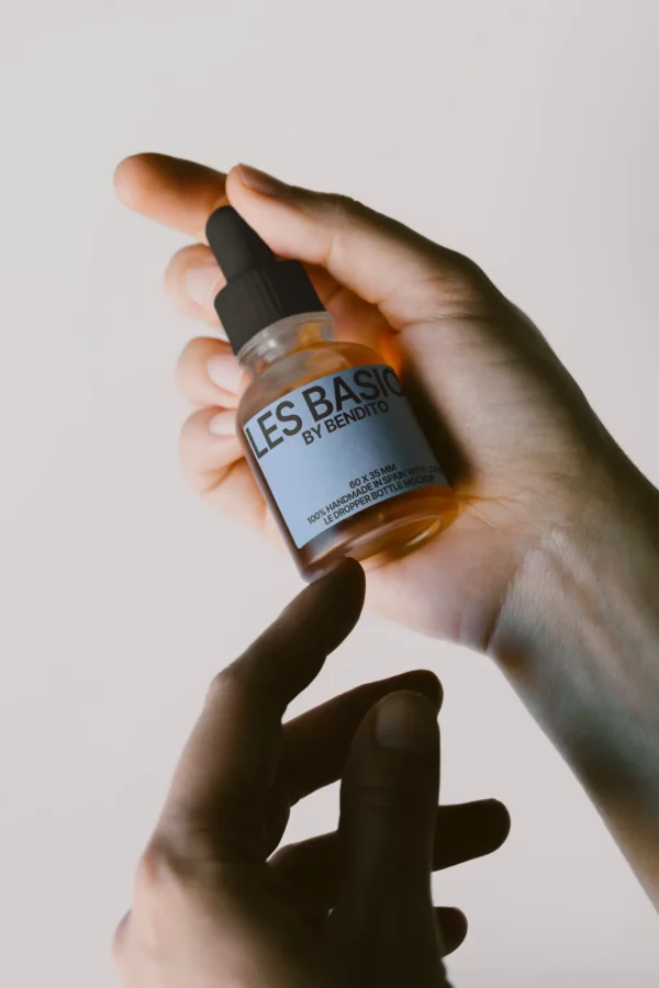 Dropper bottle mockup held between two hands with minimalist lighting. Features a label area of 60 × 35 mm, ideal for skincare or essential oil packaging.