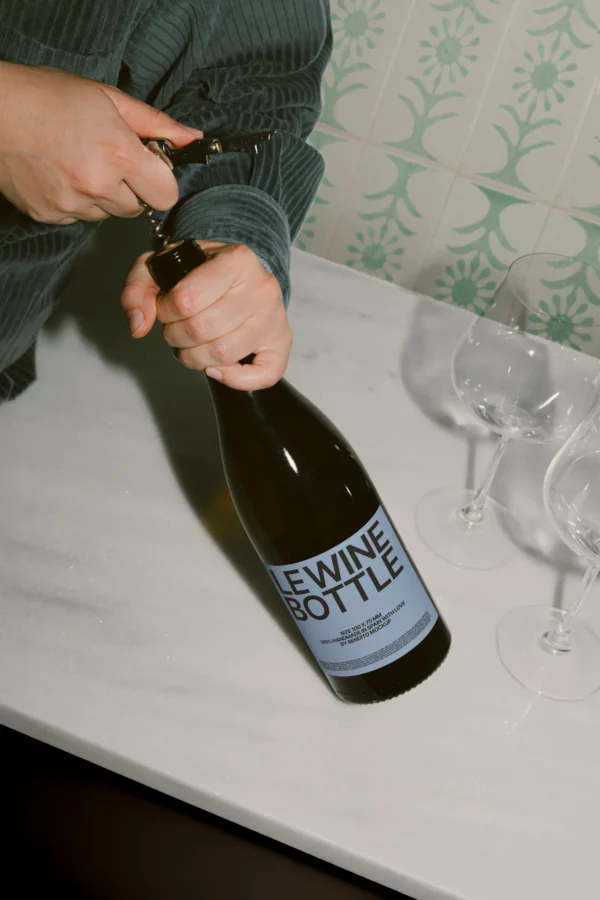 Wine bottle mockup held in hand while uncorking, surrounded by elegant glassware. Showcases a 75cl label area for beverage branding.