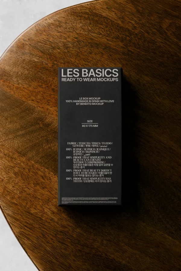 A black mockup box labeled LES BASICS READY TO WEAR MOCKUPS sits on a wooden surface. White text displays size and fabric details, while the box casts a shadow across the table.