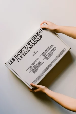Two hands hold a rectangular white box with black text that reads, “LES BASICS / BY BENDIT / LE BOX MOCKUP,” making it a perfect packaging mockup, with additional smaller text featured on the lid.