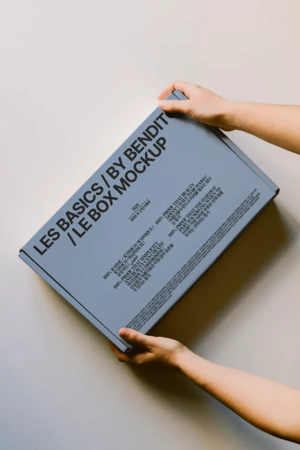 A person holds a rectangular, light gray mockup box labeled Les Basics by Bendit | Le Box Mockup with additional text, positioned against a plain, light-colored background.