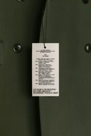 A close-up mockup of an olive green coat with black buttons, showing a white tag hanging from a black string, listing fabric composition and care instructions in black text.