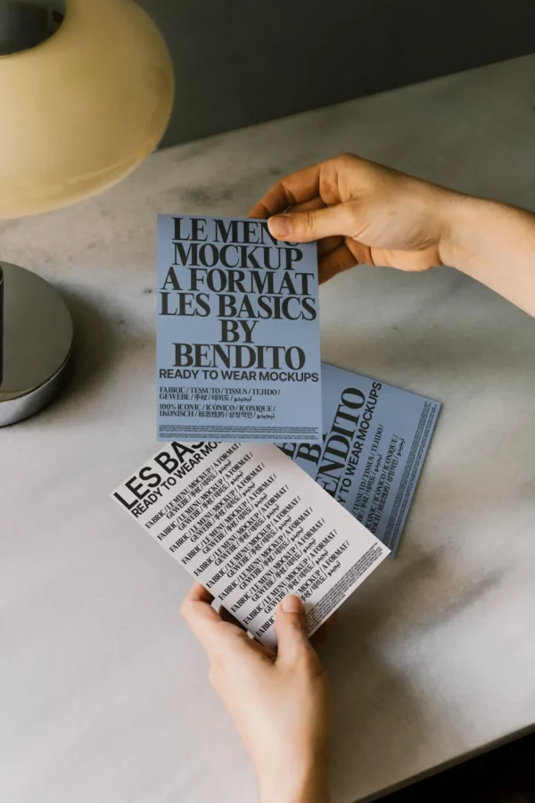 A person holds three blue and white mockup flyers with large, bold, black text on a light-colored table next to a lamp. The cards showcase various design and typography samples.