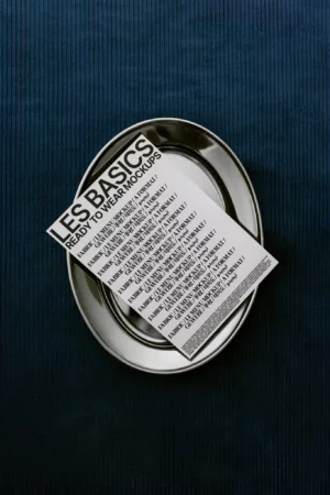 A small white mockup card with black text titled LES BASICS READY TO WEAR MOCKUPS rests on two stacked silver plates, set on a dark blue textured surface.