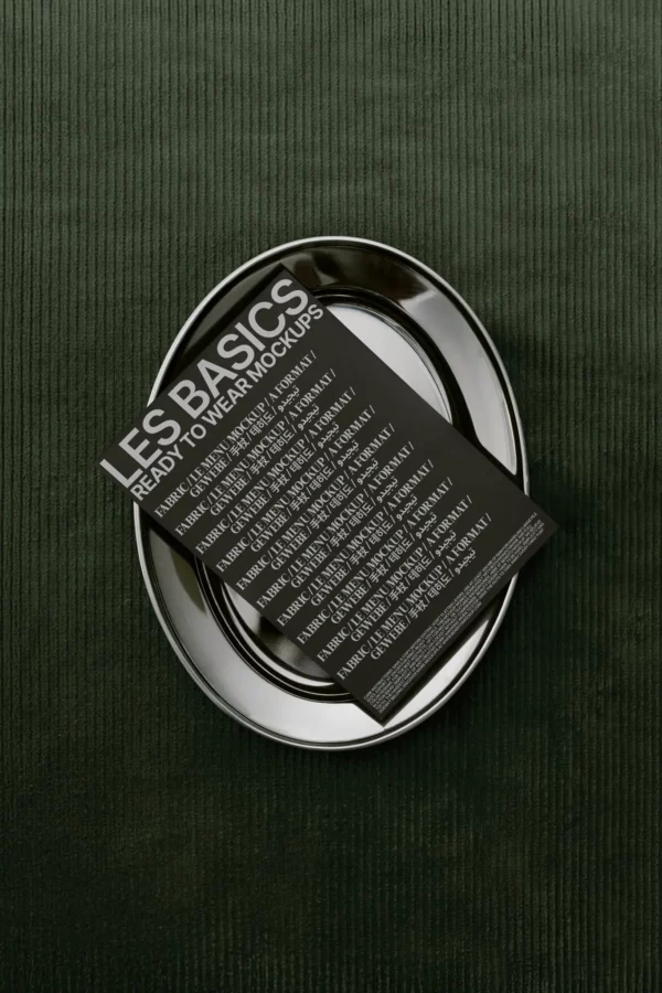 A black mockup card with white text titled LES BASICS READY TO WEAR MOCKUPS rests on stacked black plates atop a dark green textured surface.