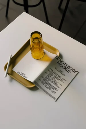 A yellow tray with a white pen, a stacked yellow glass, and an open notebook displaying the text “LE NOTEBOOK MOCKUP” create a perfect setting for showcasing your next mockup on a white table.