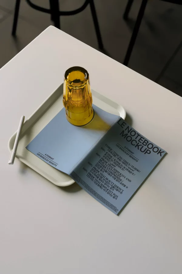 A white tray holds an open blue notebook labeled “Notebook Mockup,” a white pen, and an upside-down yellow glass, all arranged on a white table with another dark chair visible in the background.