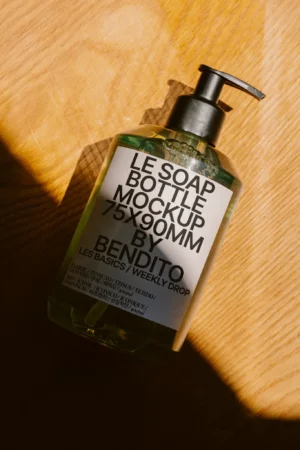 A clear pump soap bottle mockup labeled LE SOAP BOTTLE MOCKUP 75X90MM BY BENDITO lies on a wooden surface, casting a shadow in sunlight.