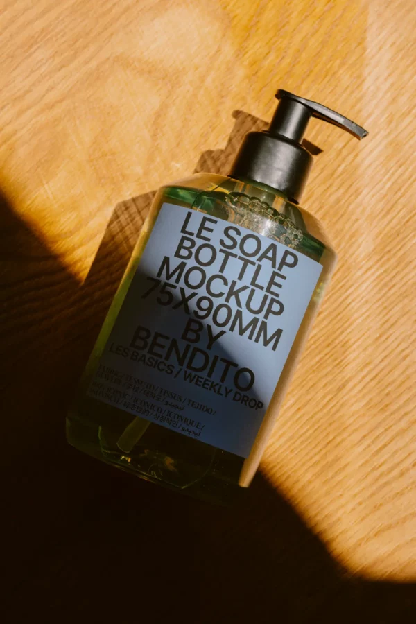 A clear pump soap bottle mockup labeled LE SOAP BOTTLE MOCKUP 75X90MM BY BENDITO lies on a wooden surface, dappled with partial sunlight and shadow.