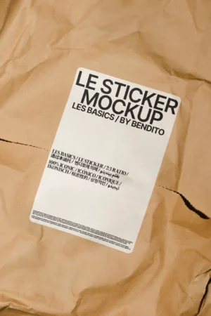 A rectangular white mockup sticker with black text reading LE STICKER MOCKUP LES BASICS / BY BENDITO is placed on crumpled brown paper. The sticker also features smaller text in various languages.