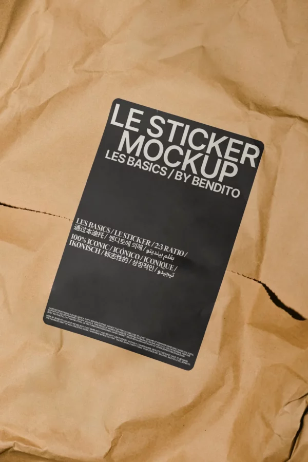 Rectangular black mockup sticker with white text reading LE STICKER MOCKUP LES BASICS / BY BENDITO, placed on crumpled brown paper. Smaller text and details appear below the main title on the sticker.