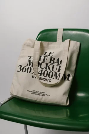 A cream-colored tote bag mockup with bold black text rests on a green plastic chair. The text on the bag includes the words LE TOTE BAG MOCKUP 360 x 400 MM BY RENDITO.