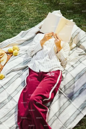 A stock photo shows a person lying on a striped blanket on grass, dressed in a white striped shirt and red pants, holding an open book over their face. Apples and bread are arranged beside them on the blanket.