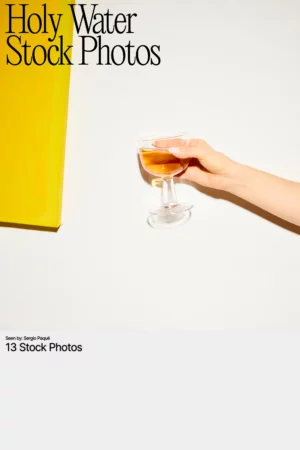 Stock image featuring a hand holding a glass of amber liquid against a white wall with a bold yellow canvas, used as the cover for the Holy Water stock photo collection.