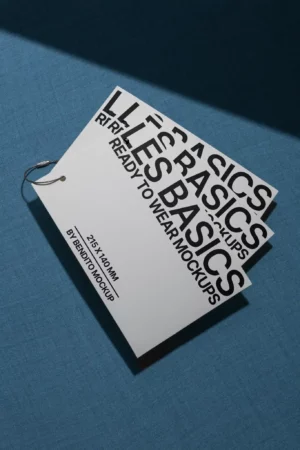 A white card with bold black text reading BASICS READY TO WEAR MOCKUPS is attached to a metal ring, resting on a blue textured surface. Shadows cross the scene, creating a minimalist and modern look.
