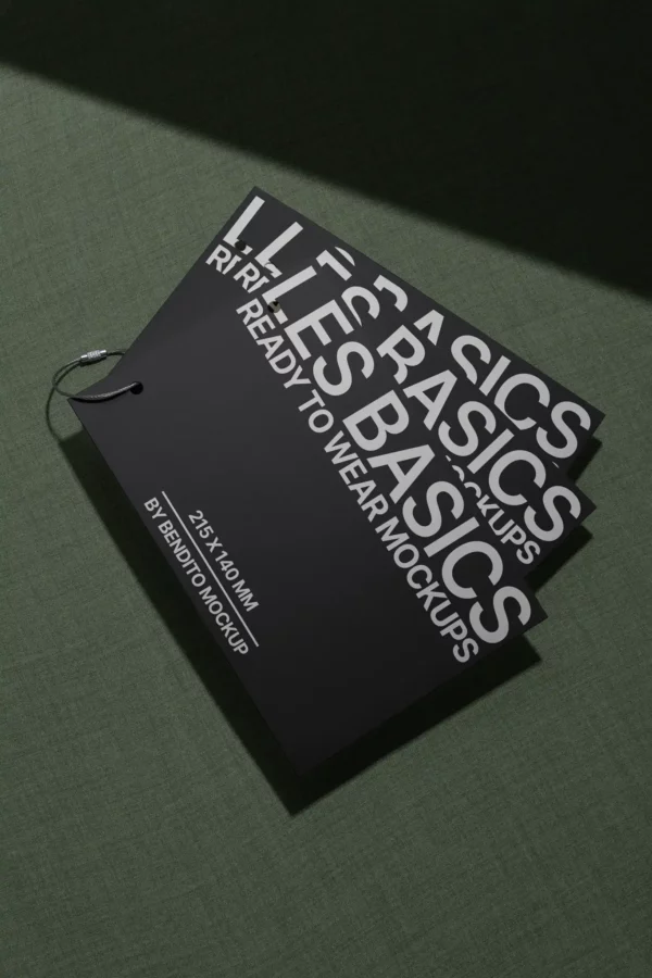 A set of three black rectangular tags labeled BASICS and READY TO WEAR MOCKUPS in bold white text, connected by a metal ring, placed on a green textured surface in dramatic lighting.
