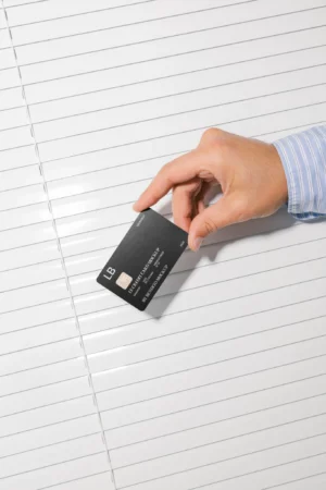 A hand in a blue-striped shirt holds a black credit card over a white, horizontally-slatted surface.