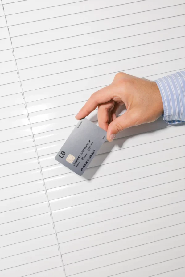 A hand in a blue striped shirt holds a gray credit card, about to slip it between the slats of closed white window blinds.