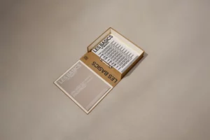A beige box labeled LES BASICS is open on a neutral surface, revealing a booklet or card inside with bold black text and a minimalist design. The lid of the box displays the same LES BASICS branding.