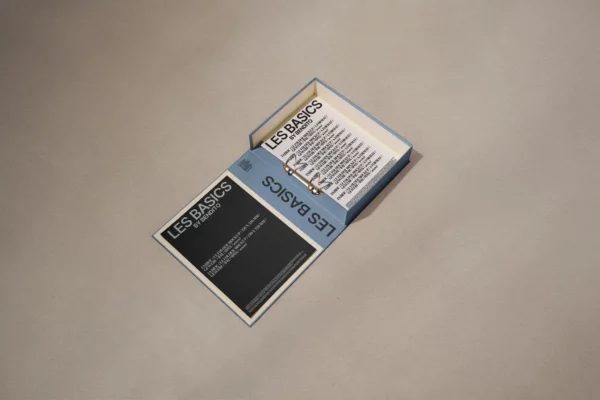 An open box labeled LES BASICS contains a ring-bound stack of cards with text, positioned on a plain, beige surface. The box lid displays the same LES BASICS title and additional information.