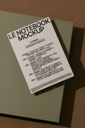 A white notebook titled LE NOTEBOOK MOCKUP rests on a closed greenish-gray folder, displayed on a brown textured surface. The notebook cover features bold black text in multiple languages.
